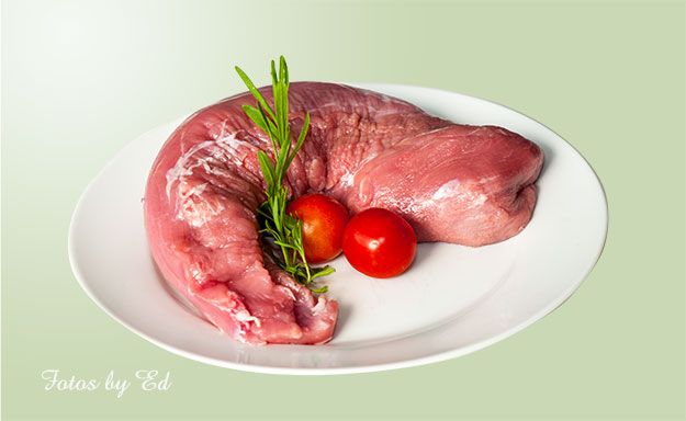 Ein rohes Schweinefilet, kunstvoll arrangiert auf einem Teller mit Rosmarinzweig und Kirschtomaten vor einem grünen Hintergrund.