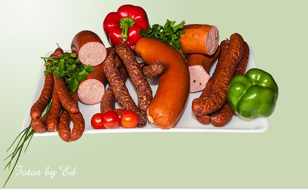 Ein Tablett mit verschiedenen geräucherten und gekochten Würsten, darunter Krakauer, Mettwürste und Landjäger, ergänzt durch rote und grüne Paprikaschoten, Kirschtomaten und frische Kräuter.
