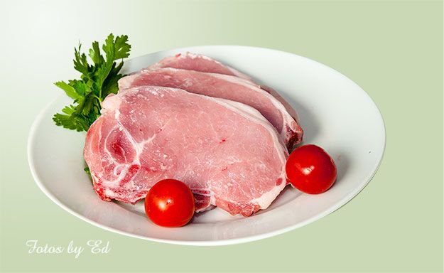 Ein weißer Teller mit drei rohen Schweinekoteletts, garniert mit zwei roten Kirschtomaten und einem Zweig frischer Petersilie.