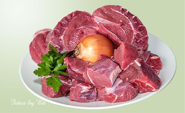 Frisches Gulaschfleisch mit Zwiebel und Petersilie auf einem weißen Teller angerichtet.