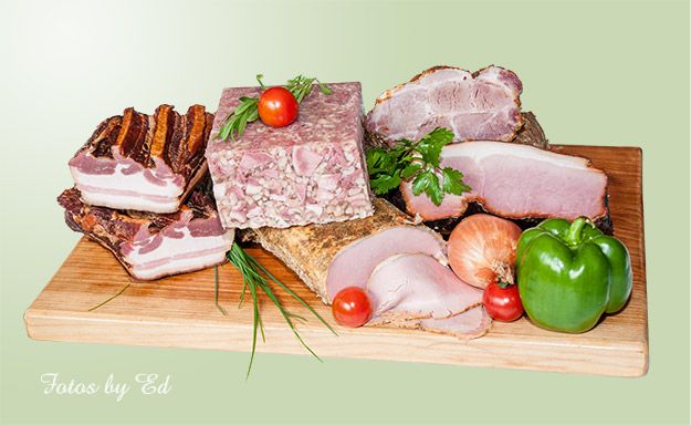 Verschiedene geselchte Fleischwaren, inklusive Speck und Schinken, auf einem Holzbrett mit Gemüse dekoriert.