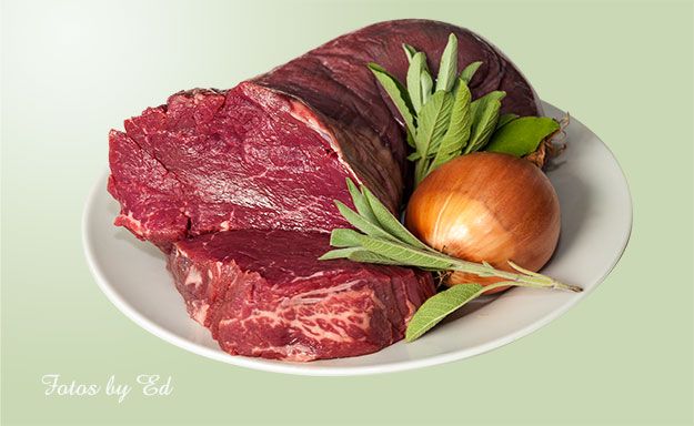 Ein Teller mit einem großen Stück rohem Rindfleisch, das teilweise aufgeschnitten ist. Daneben liegen eine ganze Zwiebel und einige frische Kräuter, darunter Salbei.